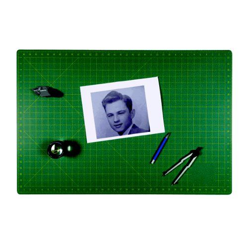 A SpeedPress 35.8" x 47.6" Magic Self-Healing Cutting Mat with a green grid design, displaying drafting tools, a utility knife, and a black-and-white photo of a man placed on its surface.